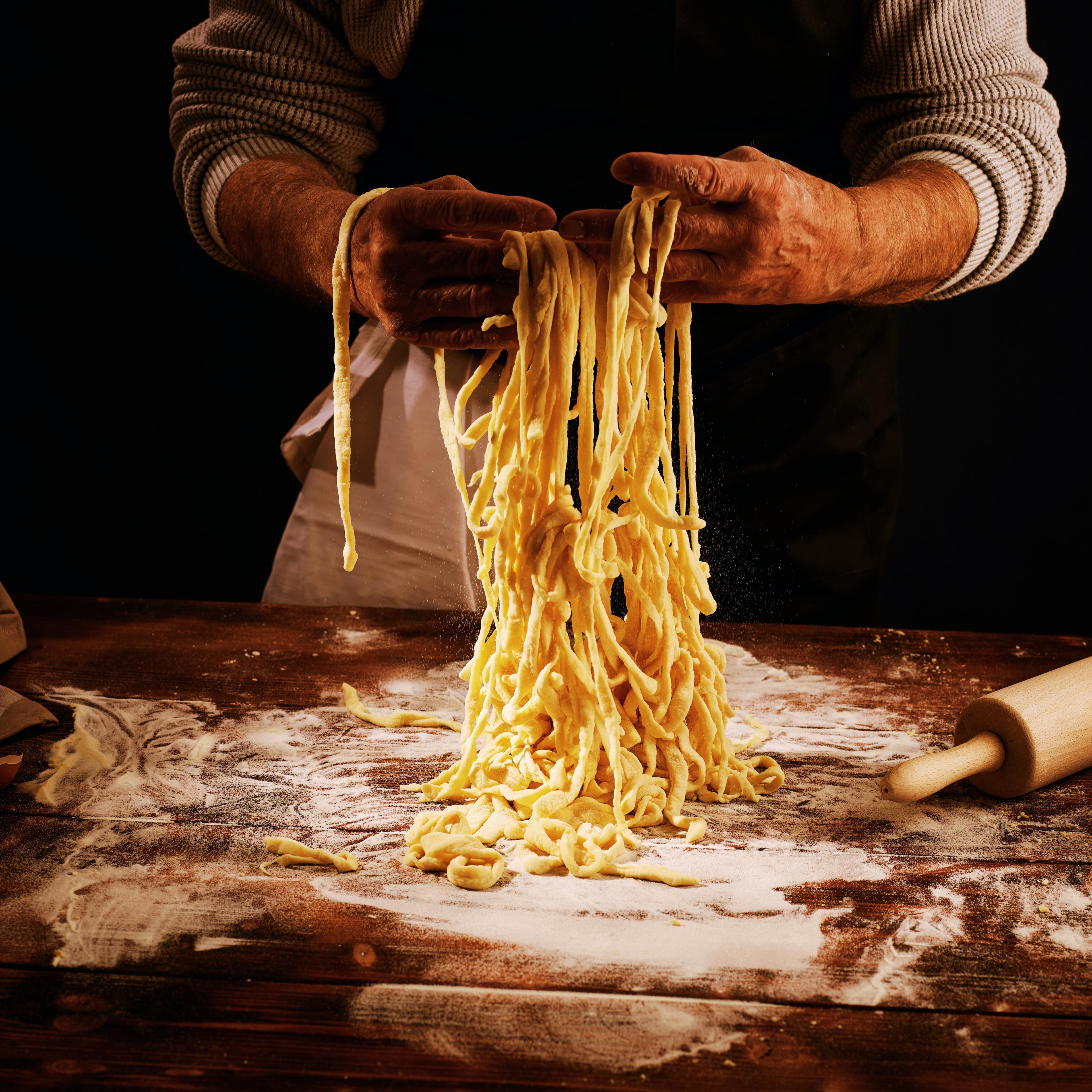 Hands lifting freshly cut pasta from a floured board, a rolling pin beside it