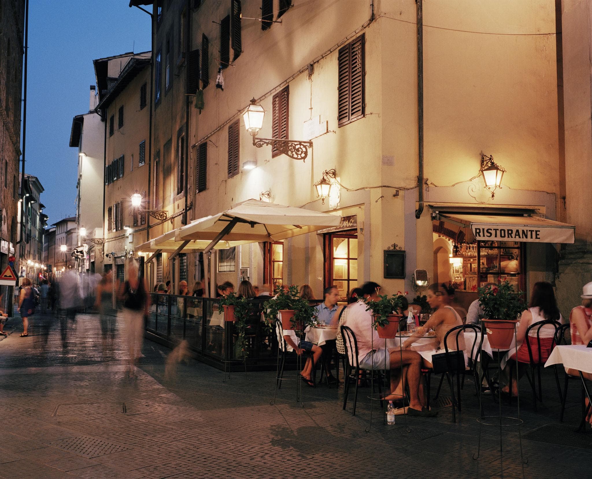 A restaurant terrace on a lamplit street corner at dusk, tables set out on the cobbles