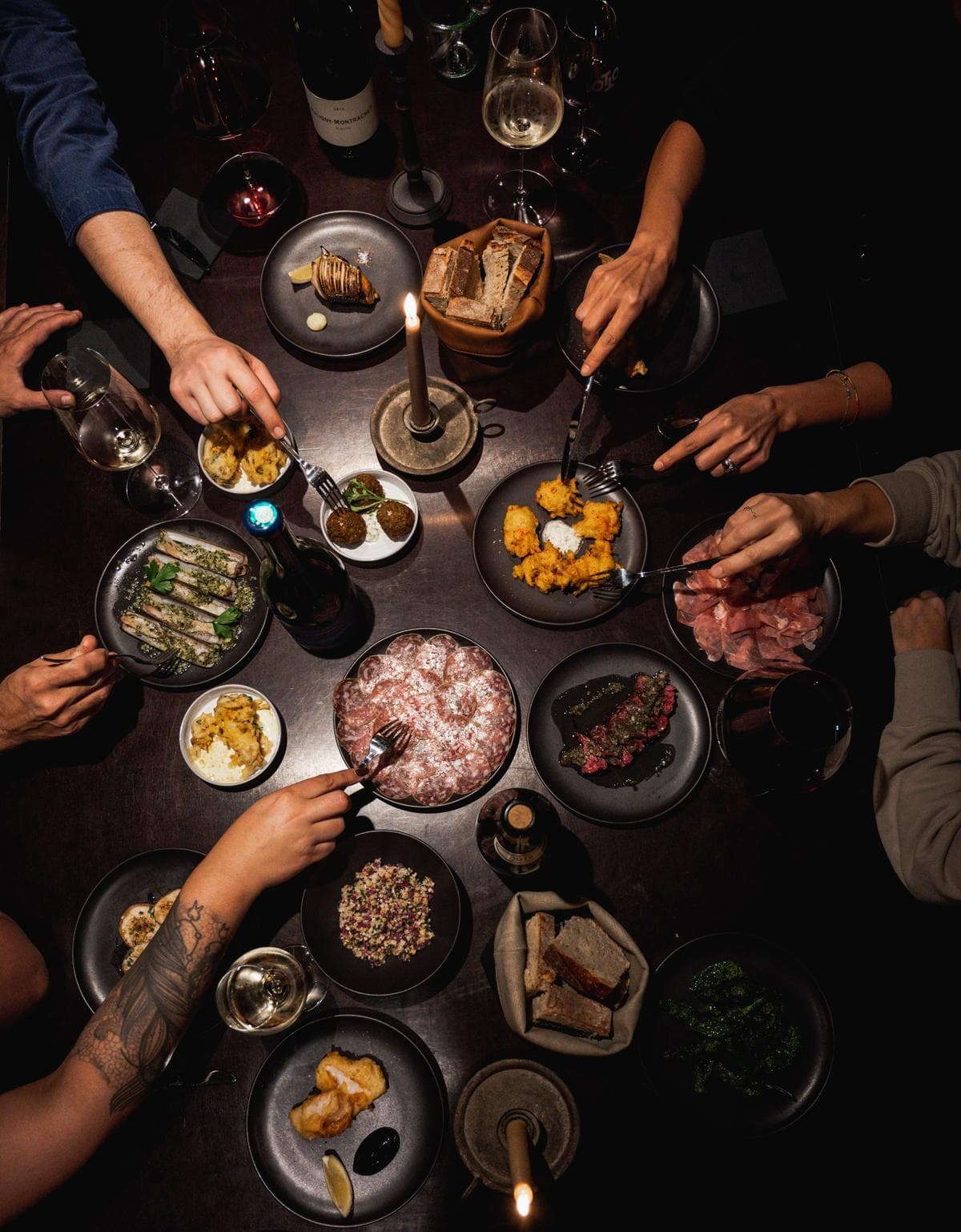 Looking down on a shared table mid-meal, hands reaching across plates by candlelight