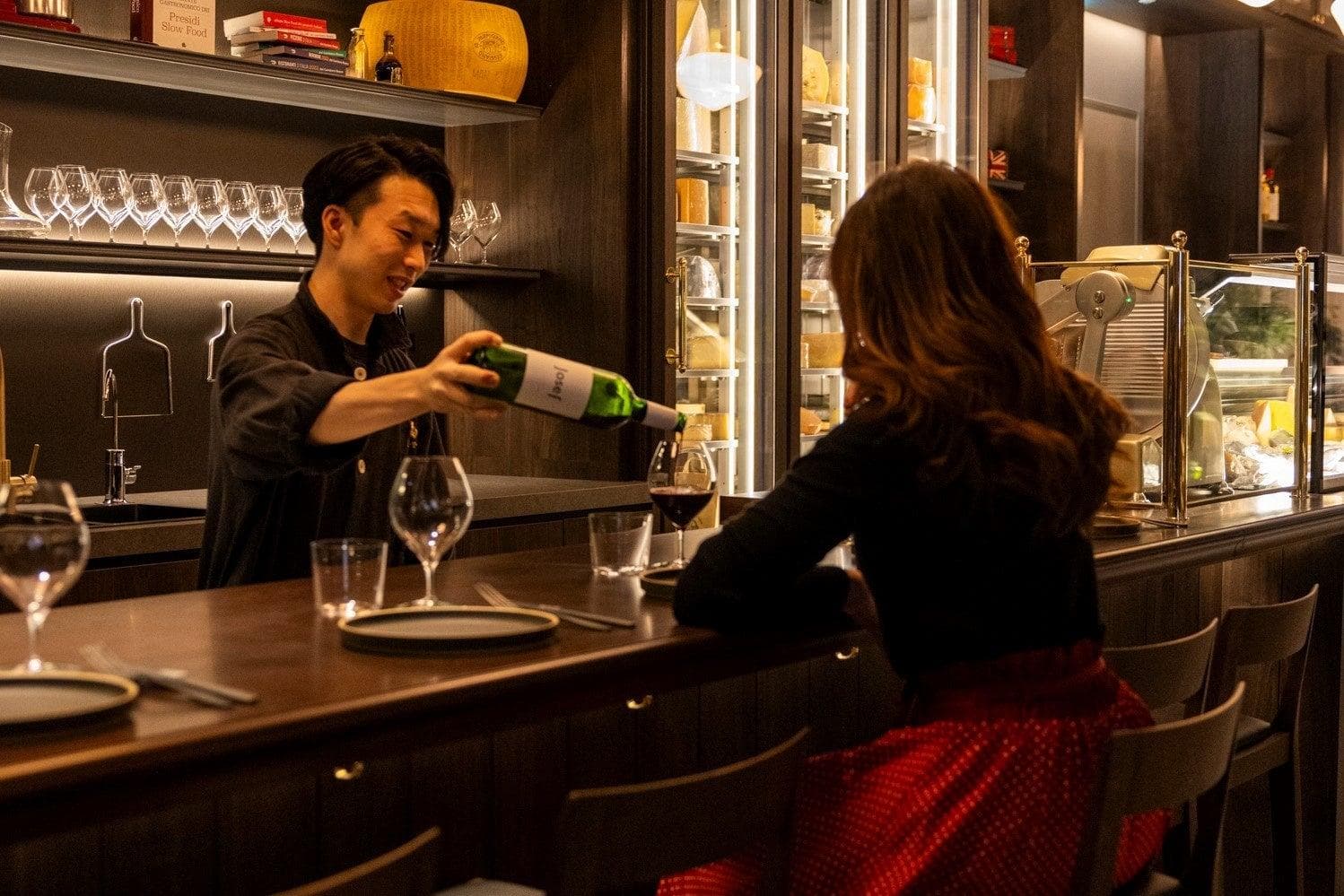 A glass of wine poured for a guest at the bar, the cheese cabinet lit behind