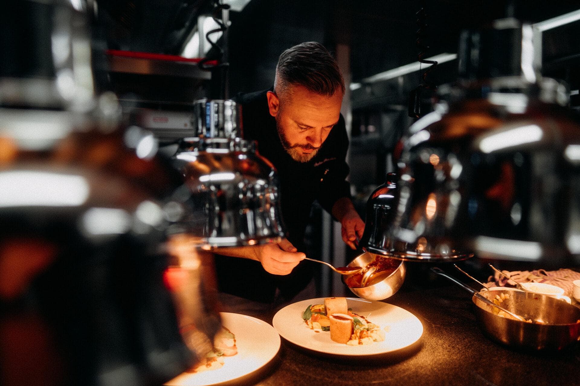 A chef spooning sauce over plates at the pass, under the heat lamps