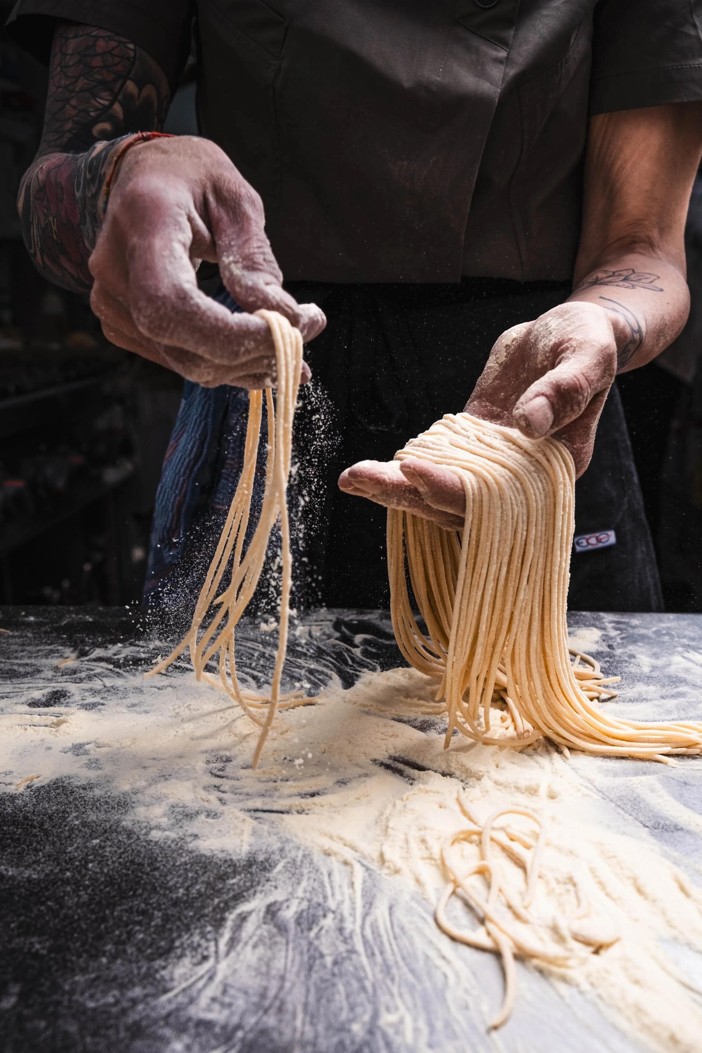 A cook shaking flour from freshly cut spaghetti over the bench